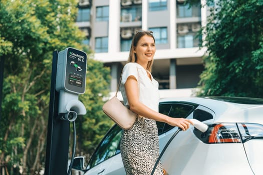 Young woman travel with EV electric car charging in green sustainable city outdoor garden in summer. Urban sustainability lifestyle by green clean rechargeable energy of electric BEV vehicle innards