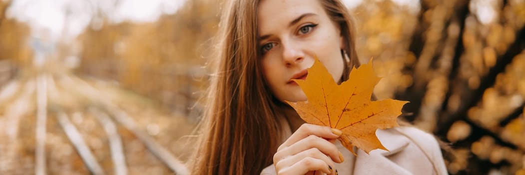 Portrait of a woman with an autumn maple leaf. Railway, autumn leaves, a young long-haired woman in a light coat coat, close-up.