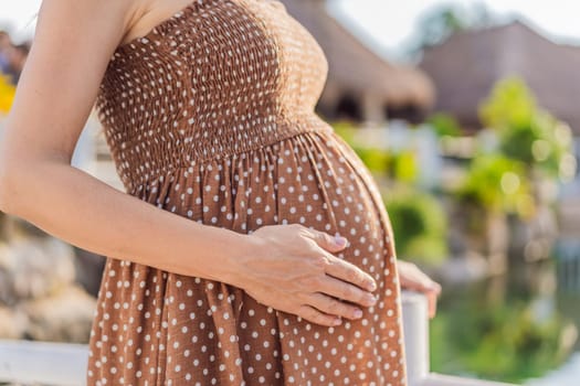 Pregnant woman hugging her tummy standing outdoors surrounded by nature. Pregnancy, expectation, motherhood concept.