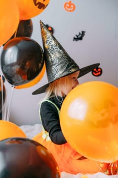 Children's Halloween - a girl in a witch hat and a carnival costume with airy orange and black balloons at home. Ready to celebrate Halloween.