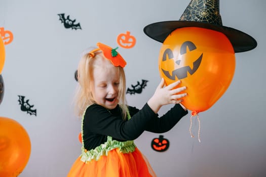 Children's Halloween - a girl in a witch hat and a carnival costume with airy orange and black balloons at home. Ready to celebrate Halloween.