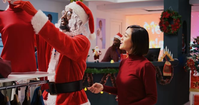 Manager wearing Santa Claus costume around shopping mall fashion boutique chatting with elderly woman. Supervisor in holiday themed suit showing senior client red garment piece