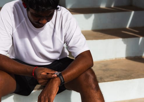 Close up Asian young athlete sport runner black man wear modern time smart watch he sitting resting before training running at the outdoor street health park, healthy exercise before workout concept