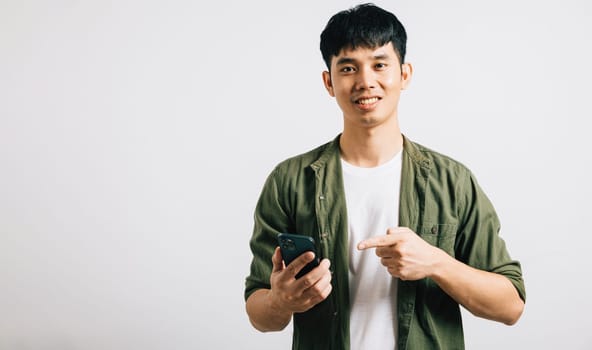 Portrait of a confident Asian man smiling while pointing at a smartphone for online shopping. Studio shot isolated on white, emphasizing smart technology usage. Copy space is present.