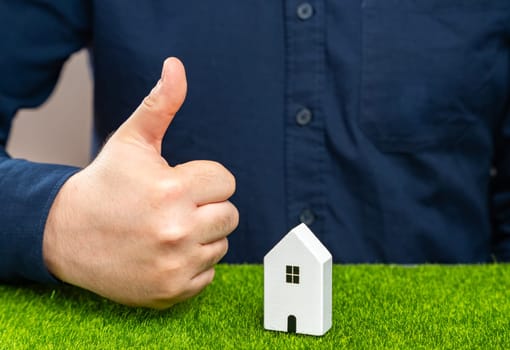 A man shows a thumbs up near a house. Successful real estate investment. Completion of a favorable deal. Approval of a mortgage or loan, making the purchase or renovation possible. Approves condition
