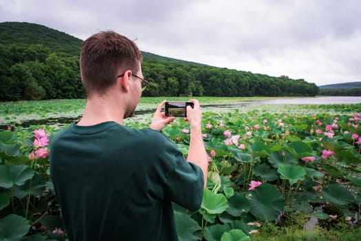 A young man with glasses photographs vibrant pink lotus flowers on a smartphone. He stands by a tranquil lake surrounded by lush greenery in the Russian Far East