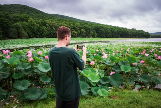 A young Russian man is capturing a scenic view of Komarov lotuses on his smartphone at a lake in the Russian Far East, highlighting modern technology and nature