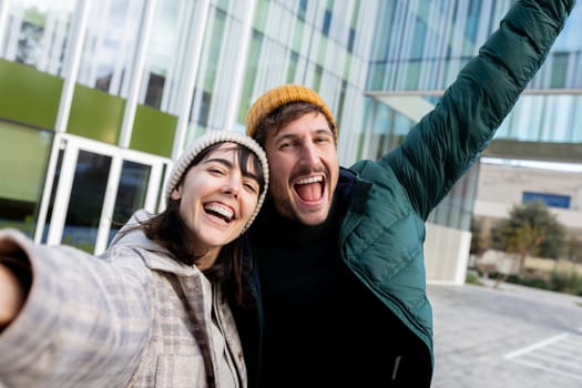 Excited young couple taking selfie outside office building, celebrating success looking at camera