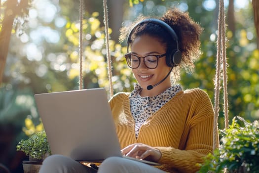 A woman wearing glasses and a headset is sitting on a swing and using a laptop. She is smiling and she is enjoying her work