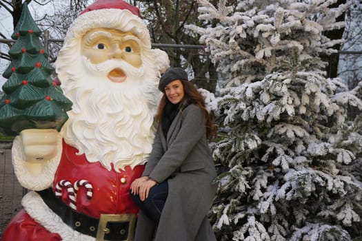 A cheerful woman in a gray coat and beret leans next to a Santa statue and a snow-covered tree, enjoying holiday festivities
