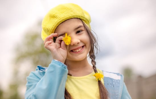 Portrait of a funny charming little girl with two pigtails, in a yellow knitted beret, a blue raincoat, holding a dandelion flower near her eyes, stands in nature in the spring, smiling