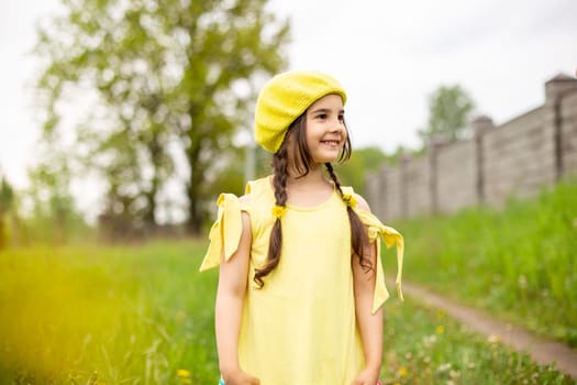 happy charming girl in a yellow knitted beret and a yellow dress, with two pigtails stands in nature, green grass with yellow dandelions, in the spring, looks to the left