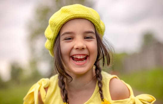 Outdoor shot portrait of a little adorable happy girl in a yellow knitted beret and yellow dress looking straight into the camera and laughing. Happy childhood concept.