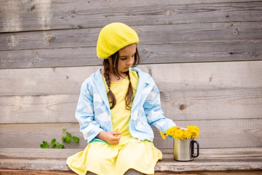Adorable little girl with two pigtails, in a yellow beret, blue raincoat, sits in the countryside near a old wooden building with bouquet of dandelions in a mug