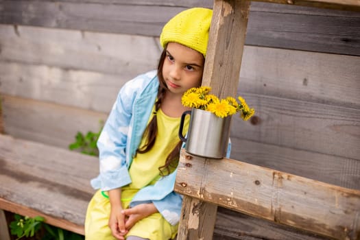 Portrait of a sad adorable little girl with two pigtails, in a yellow beret, blue raincoat, sits in the countryside near a old wooden building with a ladder and a bouquet of dandelions in a mug