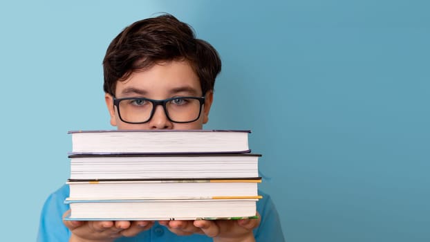 A teenage boy with glasses peeks out from behind a stack of books, on a blue background.Copy space