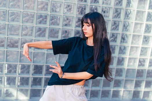 A dancer showcases her unique style with graceful movements near a glass brick wall. Her black top contrasts with the light background, highlighting her focus and artistry.