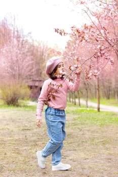 A little girl in a pink jumper and blue jeans, sniffs a branch of cherry blossoms, in the park during the day. Vertical