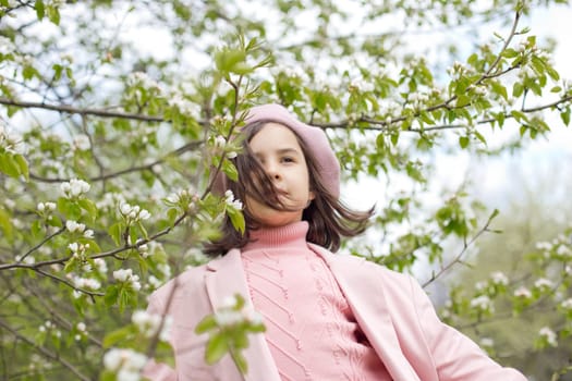 Portrait of a beautiful brunette in a pink jumper and beret stands on a blooming white apple tree in the park, with her hair flowing. Close-up