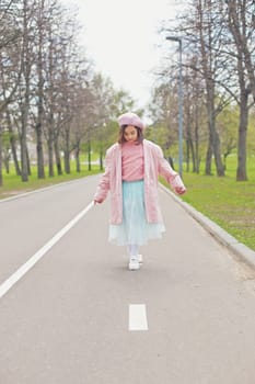 a little charming girl in a pink jumper, jacket, beret and light blue skirt walks along an asphalt road in the park. Vertical