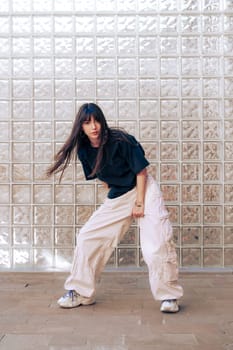 A dancer showcases her talent with a dynamic pose, wearing loose clothing and sneakers. The modern glass brick wall adds an artistic backdrop to her expressive movement.