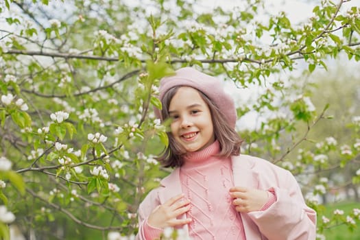 Portrait of a happy beautiful little girl in a pink jumper and beret stands next to the branches of a blossoming white apple tree in the park. Close-up