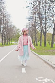 a happy little charming girl in a pink jumper, jacket, beret and light blue skirt walks along an asphalt road in the park. Vertical