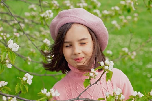 A happy little girl in a pink jumper and beret stands with her eyes closed in a blooming white apple tree in the park. Close-up