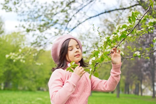 A beautiful little girl in a pink jumper holds a branch of a blossoming white apple tree in the park during the day, closing her eyes. Close-up