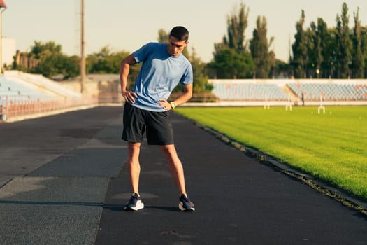A young male athlete stretches his legs on a track field surrounded by green grass and empty bleachers, preparing for a running workout during the evening light.