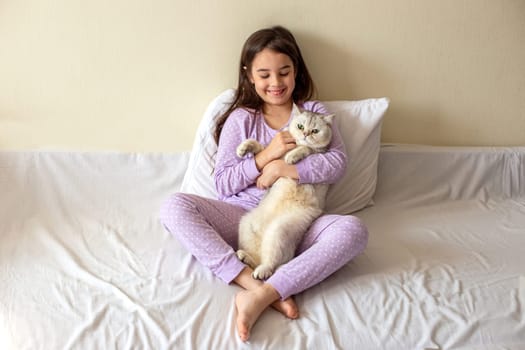 A happy little brunette girl in purple pajamas, with her dark hair loose, sits hugging a white charming cat on a white bed, smiling