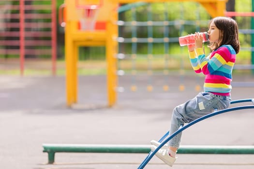 Little girl 5-6 years old, sits on the playground in summer, drinks water and a pink plastic bottle. Copy space