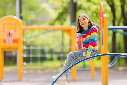Charming smiling girl 5-6 years old, sitting on the playground in summer, holding a pink plastic water bottle