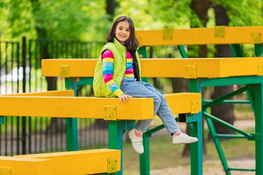 a cute smiling little girl, in a multi-colored striped jumper and a green vest, sits on a bright wooden playground, in a park, in spring