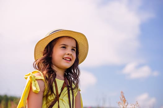 A happy cute little girl of 4-5 years old, in a straw hat and a yellow T-shirt, stands in nature in the summer, against the background of a blue sky, looks away