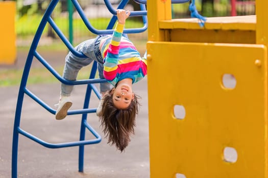 Funny little smiling girl 5-6 years old, hanging upside down on the children's stairs at the playground, in the park, in summer.Copy space