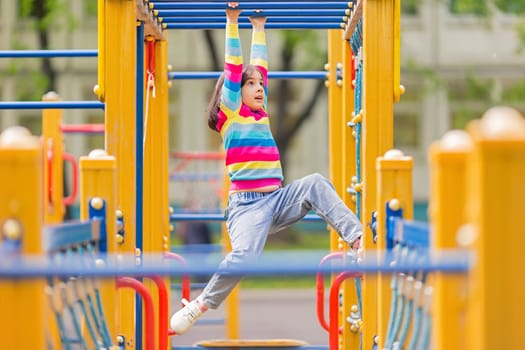 a little 5-6 years old girl hangs on a childrens ladder on a bright playground in the park in summer