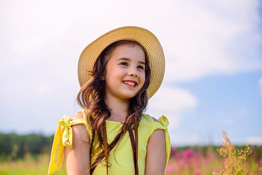 A smiling cute little girl in a straw hat and a yellow T-shirt, stands in nature in the summer, against the background of a blue sky, looks away