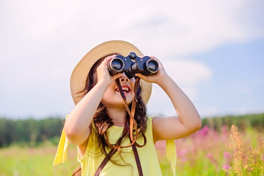 A happy cute girl 4-5 years old, in a straw hat and a yellow T-shirt, stands in a field with high grass against the blue sky in the summer, looks into the sky through binoculars. copy space