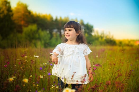 A little girl in a white dress stands on a blooming field and looking away.