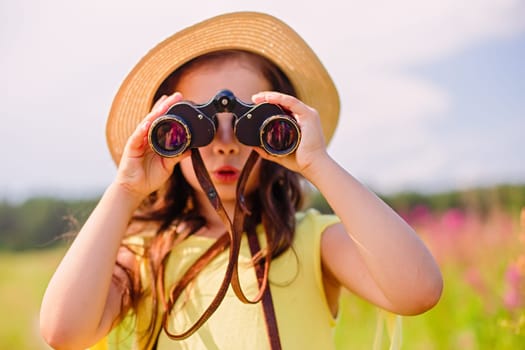 Surprised cute girl 4-5 years old in a straw hat and a yellow t-shirt in the summer stands in a field with tall grass against the blue sky, looks through binoculars. Close up