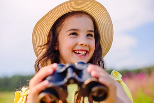 Happy cute little girl in a straw hat, stands in nature in the summer, against the blue sky, holds binoculars, looks away. Close up