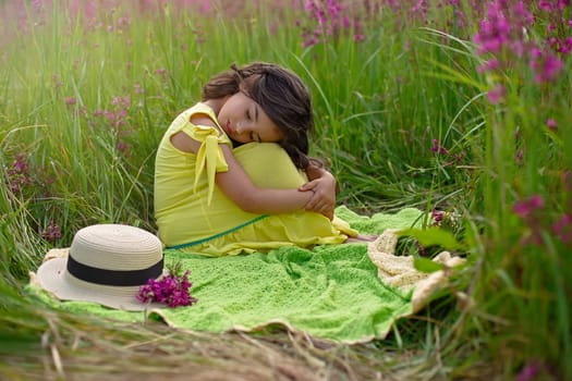 A beautiful little girl with long dark hair sits on a green knitted blanket, hugging her knees and closing her eyes, in a field of burgundy viscaria flowers in summer. copy space