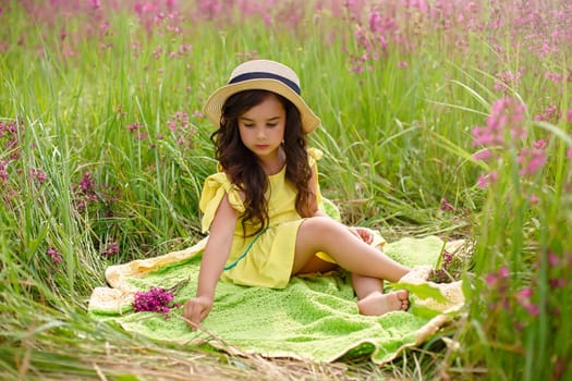 Pretty little girl 4-5 years old, in a straw hat and a yellow dress, sits on a green knitted plaid, in summer, in a field with grass and burgundy viscaria flowers. Copy space