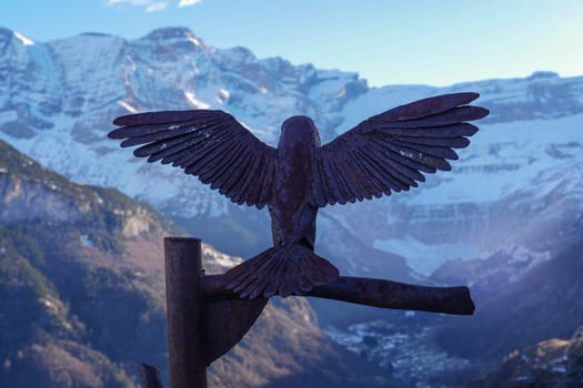 A detailed metal sculpture of a bird with spread wings stands on a branch. Dramatic mountains and snow-capped peaks rise in the background under a sunset sky.