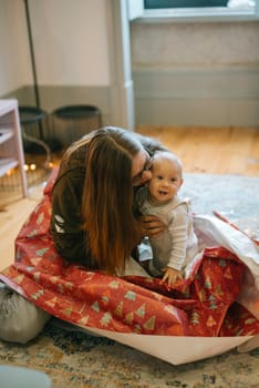 A mother joyfully interacts with her baby, both enveloped in a cheerful, festive sea of vibrant wrapping paper