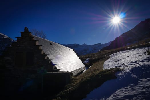 A rustic mountain hut rests on a lush hillside blanketed with snow, illuminated by a radiant sun in the clear blue sky. The scene captures the tranquil beauty of nature in the highlands.