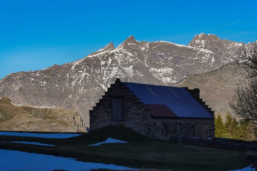 An impressive old stone structure stands quietly against majestic mountains with snow caps, surrounded by a tranquil landscape under a clear blue sky in the early morning.