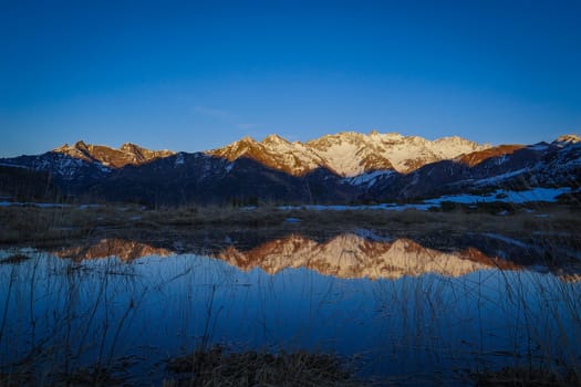 Snow-capped peaks are illuminated by the setting sun, casting a beautiful reflection in the serene water below. Golden light contrasts with the blue sky and snow.
