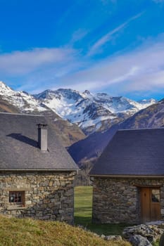 Two rustic stone cottages sit in a tranquil valley with snowy mountains rising majestically in the background under a bright blue sky, creating a serene escape for visitors.